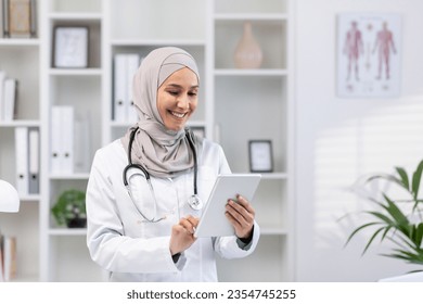 Young beautiful female doctor in hijab using tablet computer, doctor worker in white medical coat and stethoscope inside clinic office at workplace, smiling muslim woman using online application. - Powered by Shutterstock