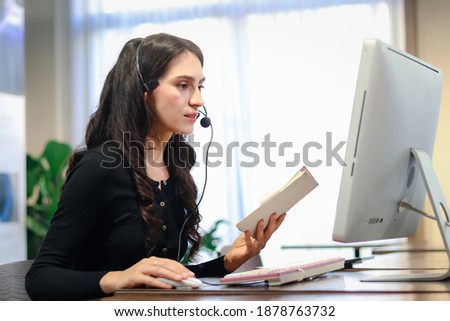 Young beautiful female with headphones working at call center service desk consultant, talking with customer on hands-free phone, woman customer support agent with headset and computer working