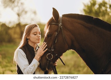 Young beautiful female jockey strokes and hugs the horse's head and prepares for the competition. Jumping training in the meadow in summer evening. - Powered by Shutterstock