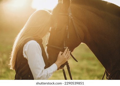 Young beautiful female jockey strokes and hugs the horse's head and prepares for the competition. Jumping training in the meadow in summer evening. - Powered by Shutterstock