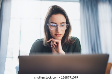 Молодая красивая женщина-модель сидит в удобном кресле и использует ноутбук. Рабочий процесс в коворкинг-студии.Панорамные окна на размытом фоне