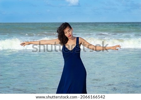 Young and beautiful female model with windy brown hair in blue dress with sea and sky in the background with open arms inspiring peace and happiness