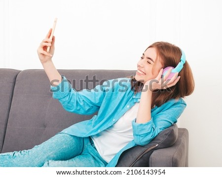 Young beautiful  female sitting at sofa. Woman lies on bed in posh apartment or hotel room in white interior in the morning. Model listening music in wireless headphones. Taking selfie photos