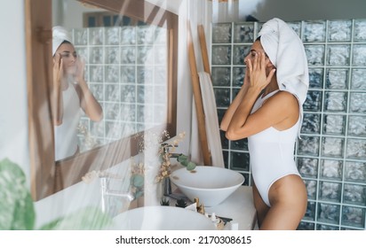 Young beautiful female with towel on head puts beauty mask on face while look to the mirror in the home bathroom. 30 years old woman doing daily morning rituals. Enjoying healthy skin care