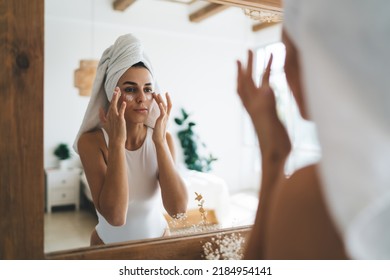 Young beautiful female with towel on head looking to mirror reflection  standing in the bathroom at home. 30 years old woman doing daily morning rituals and cleansing. Enjoying healthy skin care
