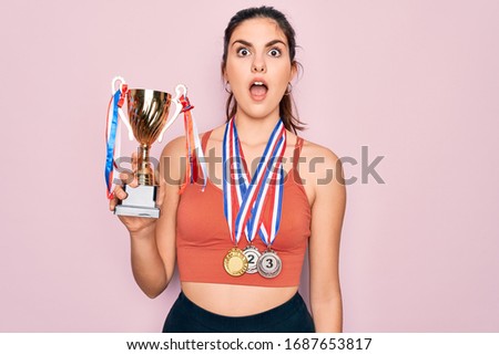 Young beautiful fitness winner athlete woman wearing sport medals and trophy scared in shock with a surprise face, afraid and excited with fear expression