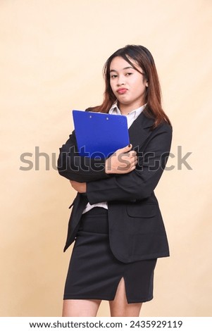 Young beautiful formal indonesia executive woman in suit frowning standing and posing hugging pen clipboard to camera, 3 by 4 body portrait on yellow background for commercial, vacancy and business
