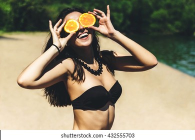 Young beautiful funny summer model holding orange in front eyes with smile and laugh on the beach in bikini sunny summer day. Vacation holiday relax concept.