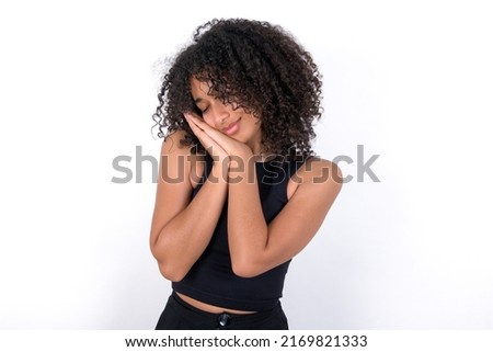 Young beautiful girl with afro hairstyle wearing black t-shirt over white background sleeping tired dreaming and posing with hands together while smiling with closed eyes.