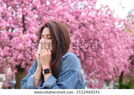 Young beautiful girl is allergic to flowers and blows her nose on a background of blooming pink sakura tree dressed in a blue denim jacket
