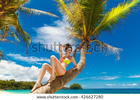 Young beautiful girl in bikini on the palm tree on a tropical beach. Tropical sky and sea in the background. Summer vacation concept.