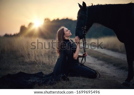 Young beautiful girl in a black cloak sits on the ground and pats her black horse on the head smiling, looks into her eyes - a concept of love between people and animals