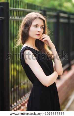 young, beautiful girl in black dress, outdoor photo on a background of steel fence, summer evening. Moddel with clean skin.