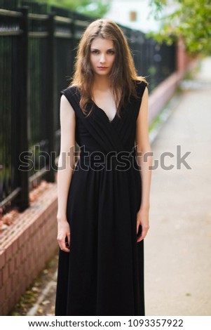 young, beautiful girl in black dress, outdoor photo on a background of steel fence, summer evening. Moddel with clean skin.