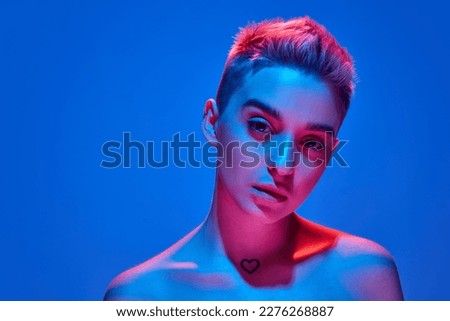 Young beautiful girl with blonde short hair posing with bare shoulders against blue studio background with pink neon light. Deep attentive look. Concept of emotions, cyberpunk style, youth culture.