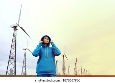 Young beautiful girl in a blue hoodie stands against the wind farm, alternative energy, sunset light
