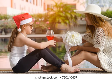 Young Beautiful Girl In Christmas Hat Holding A Glass Of Softdrink And Sitting Beside The Swimming Pool With Her Older Sister.