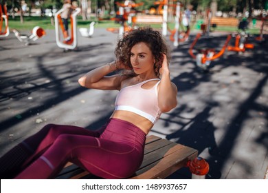 Young Beautiful Girl With Curly Hair In A Pink Sports Top And Burgundy Sports Trousers Pump Press On A Bench On The Playground In The Park