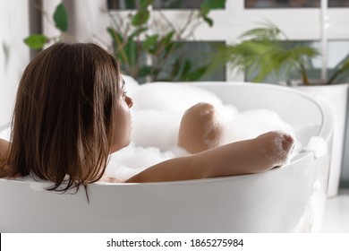 Young Beautiful Girl With Dark Hair And Tanned Skin Takes A Bath With Foam In The Interior Of A Bright Bathroom, Relaxation