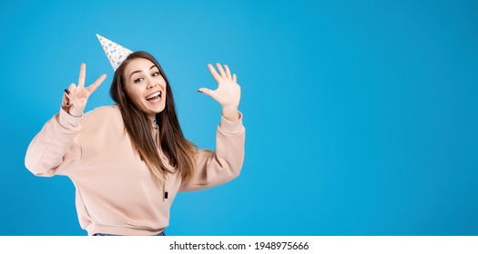 A young beautiful girl in a festive hat shows her fingers the anniversary of the age of 25. Birthday concept. Isolated on a blue background. Ultra wide image with copy space.