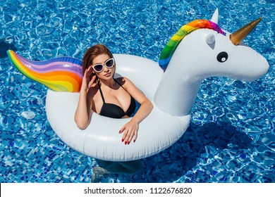 A young beautiful girl with a beautiful figure in a black swimsuit and sunglasses is resting and tanning in a swimming pool with an inflatable unicorn.