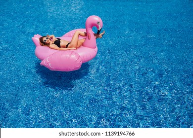 A young beautiful girl with a beautiful figure in a black swimsuit and sunglasses is resting and sunbathing in a pool with inflatable flamingos.