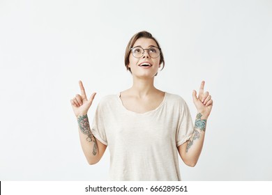 Young beautiful girl in glasses smiling pointing fingers up over white background.