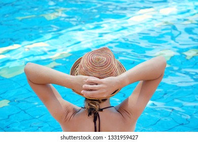 Young beautiful girl in hat relaxing in swimming pool close-up. Rear view. Young woman in hat in blue water swimming pool at resort.