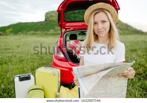 Young beautiful girl in a hat standing\
on the background of a car with a map of the area. Camping in\
nature on a summer day. Red car with Luggage for\
travel