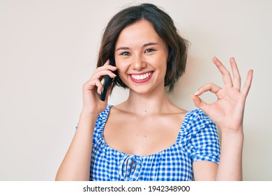 Young Beautiful Girl Having Conversation Talking On The Smartphone Doing Ok Sign With Fingers, Smiling Friendly Gesturing Excellent Symbol 