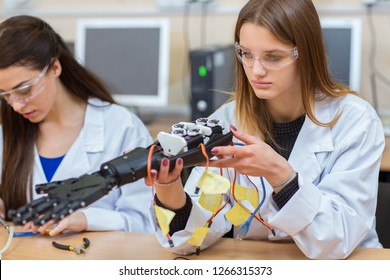 Young Beautiful Girl Holding Prototype Of Mechanical Hand In Laboratory. Student Studying Structure Of Mechanism, Verifies Whether Everything Properly Connected While Her Colleague Working Behind.