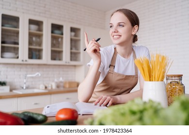 Young Beautiful Girl Housewife In The Kitchen In An Apron, Fresh Vegetables On The Table, Writes Down Her Favorite Recipes, Comes Up With Ideas For Dishes