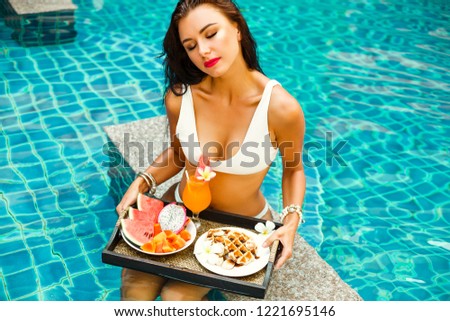 young beautiful girl lady in a white bathing suit in the pool at the hotel, in the hands of a tray with breakfast, fruit and washes. Summer holidays, holidays, smile, feeling of happiness