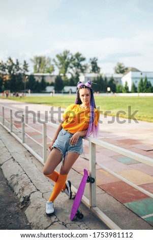 Young beautiful girl leaned on a fence in a yellow T-shirt and yellow socks, an open-air skateboard stands nearby.