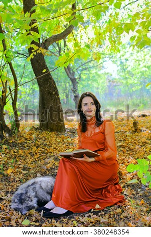 young beautiful girl with long hair in vintage red dress reading the book with her pet Raccoon Dog in the autumn forest or in the park