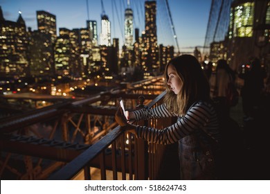 Young Beautiful Girl Looking At Mobile Phone At Brooklyn Bridge At Night. Amazing View Of New York On The Background.