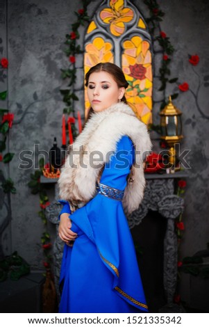 Young beautiful girl in a medieval blue dress with long sleeves and fur on her shoulders posing hands clasped together. Photo on the background of a window, fireplace and flowers.