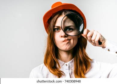young beautiful girl in an orange protective helmet looks into a magnifying glass and makes a funny face