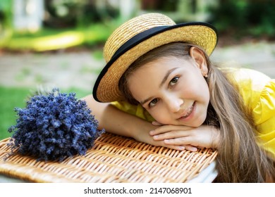 Young Beautiful Girl With Picnic Basket And Bouquet Of Flowers In Park. Closeup Portrait Of Happy Little Girl With Long Hair And Straw Hat, Looking At Camera. Beautiful Smiling Brunette Child Model. 