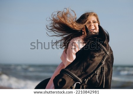A young beautiful girl in a pink dress walks and plays with a black horse on the seashore. Holiday, fun, vacation