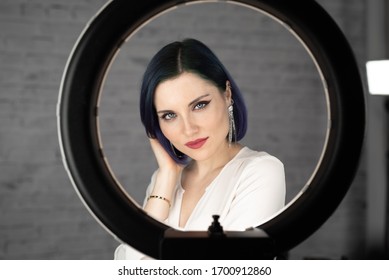 Young beautiful girl with professional makeup, blue hair and perfect skin sits in studio in front of ring light lamp posing for social media. Beauty blog. Copy space. - Shutterstock ID 1700912860
