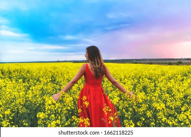 Young beautiful girl in a red dress close up in the middle of yellow field with radish flowers at sunset time
