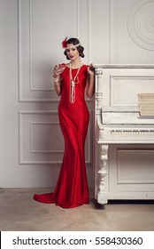 Young beautiful girl in red dress style of the 20's or 30's with glass of martini near the piano. Vintage style beautiful woman. Old fashioned makeup and retro finger wave hairstyle.