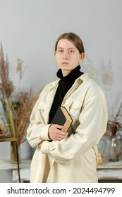 Young Beautiful Girl At School Sits In The Studio On A Chair. Girl Holding A Book In Her Hands. Portrait Photo. Attractive Girl. Props In The Background. Books On The Table