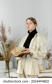 Young Beautiful Girl At School Sits In The Studio On A Chair. Girl Holding A Book In Her Hands. Portrait Photo. Attractive Girl. Props In The Background. Books On The Table