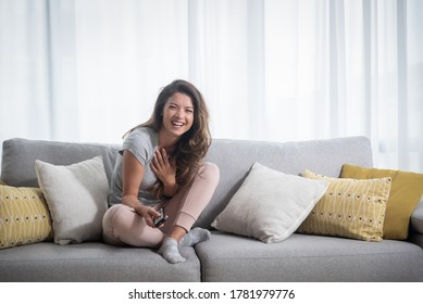 Young Beautiful Girl Sitting On Sofa Having Free Afternoon And Watching The Television Or Comedy Tv Show Movie With Big Smile On Her Face And Happy Emotion