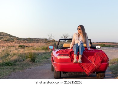 Young beautiful girl sitting on a red classic convertible car enjoying the sunset on a remote road in the countryside