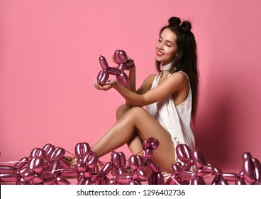 Young beautiful girl sitting in white dress hold inflatable balloon dogs happy smiling on pink background