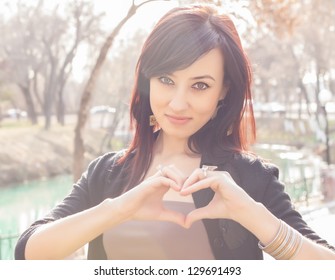 Young Beautiful Girl Smile With Hands Form Heart Shape Gesture. Photo Taken Outside, With Real Sun Light As Backlight And Real Lens Flares