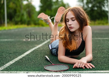 Young beautiful girl in sports top and shorts lying on the sports ground, posing. Daytime, fitness, open air, badminton, racket